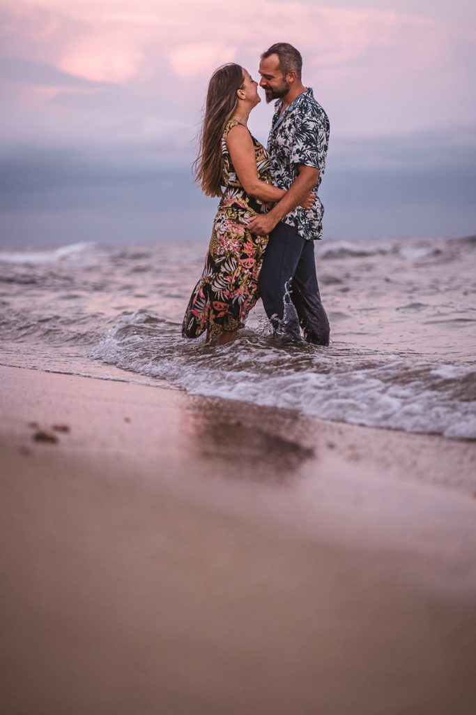 Pareja abrazándose y mirándose amorosamente mientras están de pie en el agua durante una sesión de fotos preboda al atardecer en la playa.